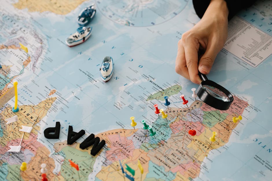 A hand using a magnifying glass over a world map with pins and toy ships